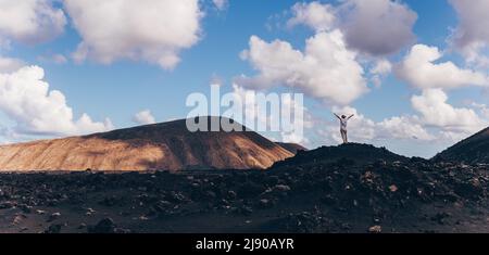 Frau, die sich mit den Händen in den Himmel erhebt und einen atemberaubenden Blick auf die vulkanische Landschaft im Timanfaya-Nationalpark auf Lanzarote, Spanien, genießt. Freiheit und Reisen Stockfoto