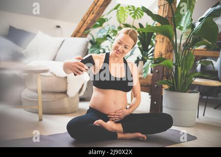 Junge glückliche und fröhliche schöne Schwangere, die Selfie mit ihrem Mobiltelefon macht und während ihres Mutterschaftsurlaubs fit, sportlich und aktiv bleibt Stockfoto