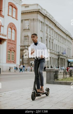 Hübscher junger Afroamerikaner mit Mobiltelefon, während er mit einem Elektroroller auf einer Straße steht Stockfoto