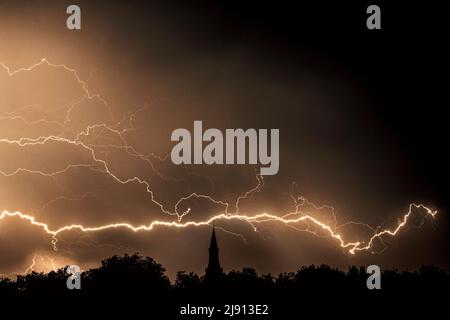 Regen fällt und Schläge von gezenkten Blitzen während Gewitter in der Nacht über Kirchturm und Bäumen Stockfoto