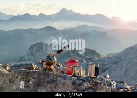 Dampfende Kaffeekocher auf dem touristischen Herd und Metallbecher auf dem Stein mit herrlichem Blick auf die Berge. Die Julischen Alpen, Soca-Tal, Slowenien Stockfoto