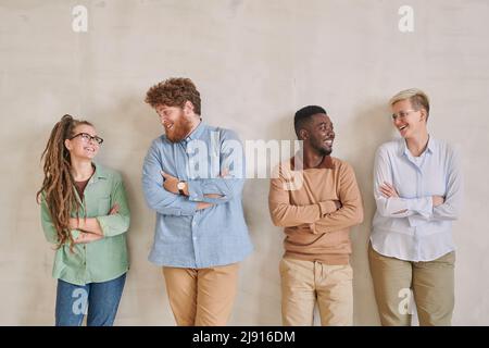 Eine Gruppe fröhlicher junger, multiethnischer Kollegen in lässigen Outfits, die in der Pause gegen die Wand stehen und miteinander plaudern Stockfoto