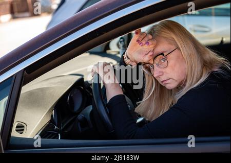 Nachdenkliche Blondine mittleren Alters in Brille sitzt hinter dem Steuer eines Autos, Blick durch das Fenster Stockfoto