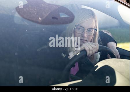 Nachdenkliche Blondine mittleren Alters in Brille sitzt hinter dem Steuer eines Autos, Blick durch die Windschutzscheibe Stockfoto