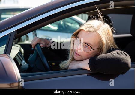 Nachdenkliche Blondine mittleren Alters in Brille sitzt hinter dem Steuer eines Autos, Blick durch das Fenster Stockfoto