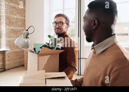 Junge, multiethnische Kollegen plaudern und tragen Pappkartons voller persönlicher Sachen, während sie in das Loft-Büro ziehen Stockfoto