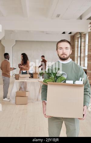 Porträt eines seriöse bärtigen Managers, der einen Karton voller persönlicher Sachen hält, während er in ein neues Büro umzieht Stockfoto