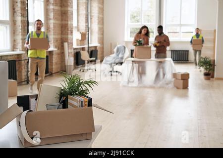 Pappkarton voller Notizblöcke und persönlicher Gegenstände, die auf den Tisch gelegt werden, Büromitarbeiter packen Kartons aus, die von Ladern getragen werden, und ziehen in ein neues Bürokonzept um Stockfoto