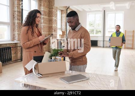 Eine junge Frau überprüft die Liste und ein schwarzer Mann zählt Notizblöcke in Kartons, während sie nach einem Umzug im Büro Sachen auspacken Stockfoto