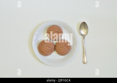 Draufsicht auf drei Kugeln Schokoladeneis auf weißem Teller mit Löffel, weißer Hintergrund mit Platz für Text Stockfoto