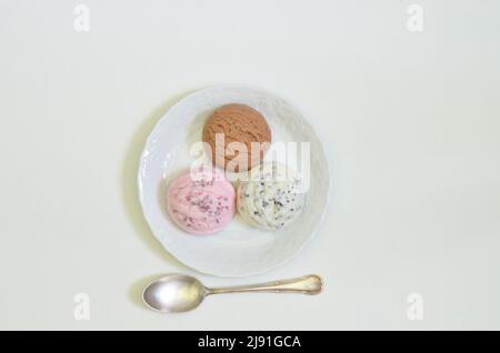 Draufsicht auf drei Kugeln Vanille-, Schokolade- und Erdbeereis auf weißem Teller mit Löffel, weißer Hintergrund mit Platz für Text Stockfoto