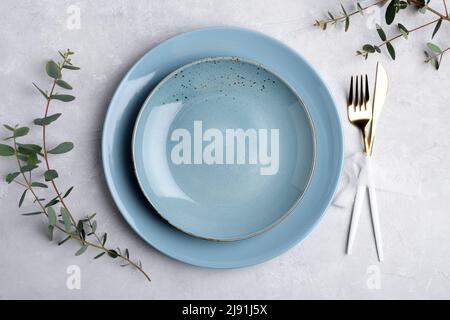 Festliche Tischkulisse mit blauer Keramikplatte, goldenem Besteck und frischen Eukalyptusblättern auf grauem Betontisch. Plattenmodup. Kopieren Sie den SPAC Stockfoto
