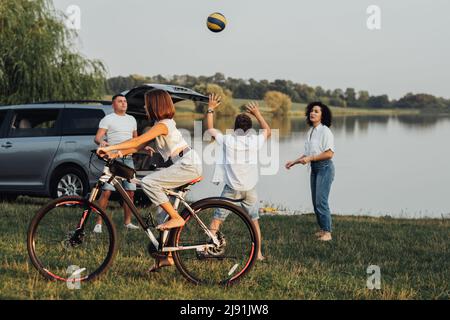 Teenager-Mädchen reiten Fahrrad, während Mutter und Vater Volleyball spielen mit ihrem Bruder im Freien am See, Happy Four Members Family genießen Stockfoto