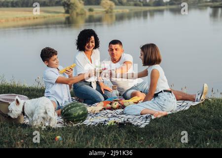 Glückliche vier Mitglieder Familie mit Hund beim Picknick bei Sonnenuntergang, fröhliche Mutter und Vater mit zwei Teenagern Kinder und Haustier genießen Wochenende im Freien Stockfoto