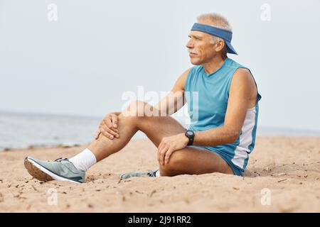 Horizontale Schuß von gealterten Mann sportliches Outfit sitzen auf Sand suchen Stockfoto