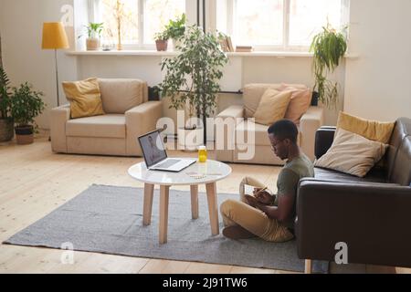 Eine lange Seitenansicht des modernen afroamerikanischen Mannes, der auf dem Boden im Wohnzimmer vor dem Laptop sitzt und einen Online-Termin mit einem Arzt hat Stockfoto