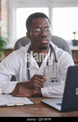 Vertikales mittleres Porträt eines modernen afroamerikanischen Hausarztes mit Brillen, der in seinem Bürozimmer mit einem Laptop sitzt Stockfoto