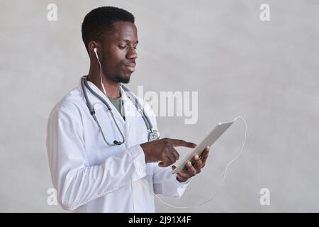 Horizontaler, mittelgroßer Schuss junger afroamerikanischer Arzt, der Kopfhörer mit digitalem Tablet trägt Stockfoto