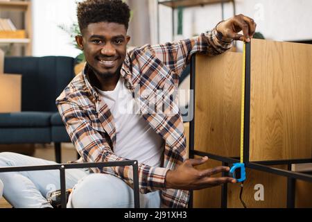 afroamerikanischer Mann in legerer Kleidung mit Maßband auf modernem Regal in gemütlicher Wohnung. Hübscher Mann, der Möbel in einer neuen Wohnung zusammengebaut hat. Stockfoto