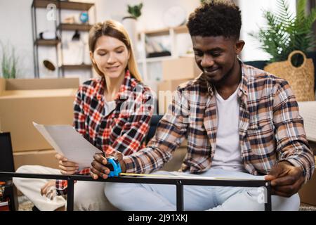 Positives multirassisches Paar in legerer Kleidung mit Hilfe von Klebeband messen Größe der neuen Möbel nach Anweisung. Konzept der Heimwerkerverbesserung. Stockfoto