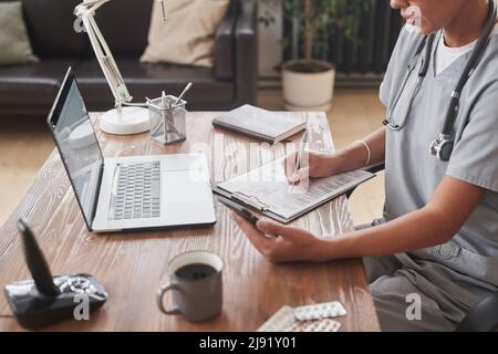 Aufnahme aus der High-Angle-Ansicht einer jungen erwachsenen medizinischen Fachärztin, die am Schreibtisch sitzt und etwas auf dem Smartphone beobachtet und Notizen macht Stockfoto