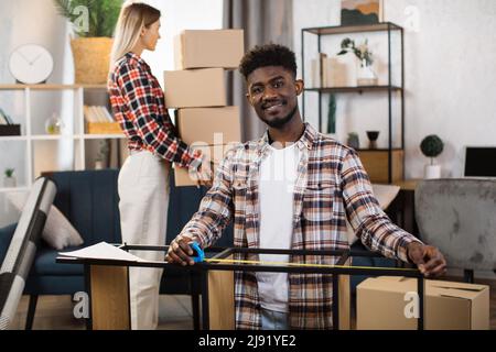 Hübscher afroamerikanischer Mann, der die Kamera anlächelt und dabei Möbel mit Klebeband misst. Attraktive kaukasische Frau trägt Haufen von Boxen auf dem Hintergrund. Bewegungskonzept. Stockfoto