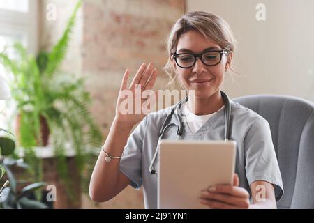 Junge Erwachsene Ärztin mit Brille am Schreibtisch sitzend, beginnt Online-Konsultation und begrüßt den Patienten mit einem Videoanruf Stockfoto
