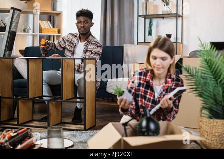 afroamerikanischer Mann, der Handbohrer für die Montage von Holzregalen verwendete, während seine kaukasische Frau Boxen auspackte, nachdem sie in eine neue Wohnung umzog. Familie und neues Kapitel im Lebenskonzept. Stockfoto