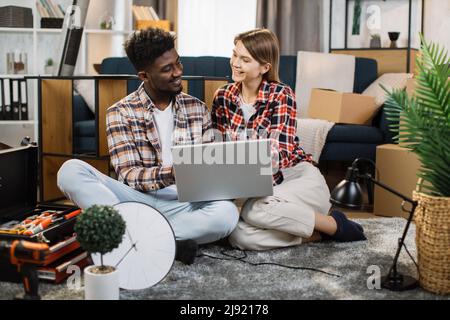 Fröhliche, multirassische Familie, die auf dem Boden mit einem Laptop sitzt und sich in einer neuen Mietwohnung sehr nett ansieht. Häusliche Lebensstile von glücklichen verheirateten Paaren. Stockfoto