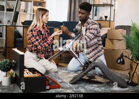 Schönes junges multiethnisches Paar mit verschiedenen Werkzeugen für die Montage von Möbeln zu Hause. Fürsorgliche Frau hilft schönen Mann Fixierung Stuhl mit Schraubendreher. Stockfoto