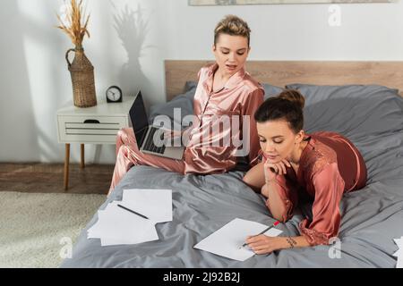 Junge großenderige Person, die auf Papier schreibt, neben dem Partner, der mit einem Laptop auf dem Bett sitzt Stockfoto