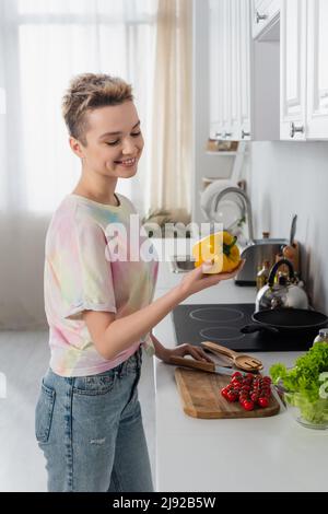 Fröhliche pansexuelle Person hält Paprika in der Nähe von frischen Kirschtomaten und Salat in der Küche Stockfoto