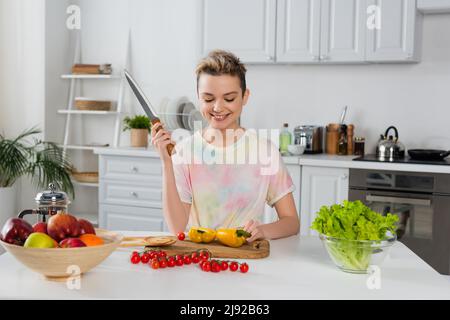 Fröhliche Bigender Person mit Messer, die in der Nähe von Paprika, Kirschtomaten, Salat und Früchten in der Küche sitzt Stockfoto