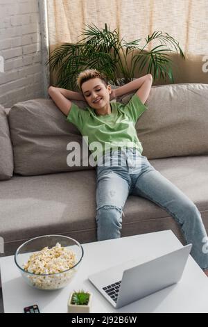 Glücklicher Großmender, der sich auf dem Sofa entspannt und sich einen Film auf dem Laptop in der Nähe einer Popcornschüssel anschaut Stockfoto