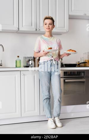 Die ganze Länge der positiven Person in Jeans hält leckere Croissants in der Küche Stockfoto