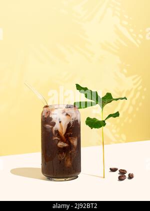 Robusta-Eiskaffee, Kaffeebohnen und Blätter auf gelbem Hintergrund. Eine Tasse Kaffee in Form einer Dose mit Milchmilch. Wiederverwendbarer Trinkhalm Stockfoto