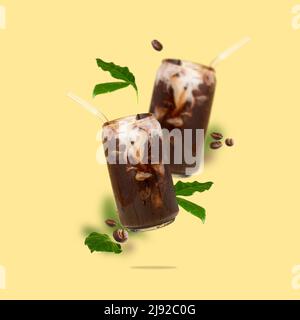 Zwei Dose-förmige Gläser mit Eiskaffee, Kaffeebohnen und Blättern in der Luft auf gelbem Hintergrund. Kalter Kaffee mit Milch. Stockfoto