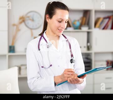 Positive junge Ärztin macht wichtige Notizen, während sie im Büro steht Stockfoto
