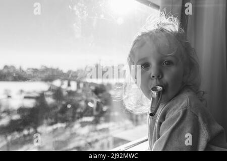 Nahaufnahme Porträt in Schwarz und Weiß eines niedlichen dreijährigen Jungen, der neben einem Fenster mit Rohr im Mund steht. Verschwommene Landschaft im Hintergrund. Stockfoto