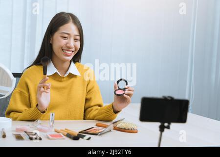 Junge asiatische Frau vloggt sich über Beauty-Produkte mit einem Video für ihren Blog über Kosmetik mit Handy-Kamera zu Hause machen. Stockfoto