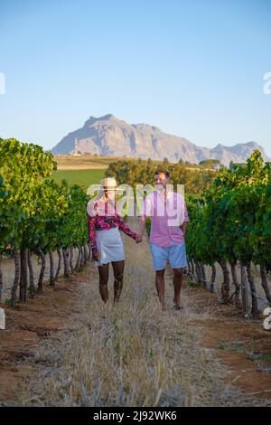 Weinbergslandschaft bei Sonnenuntergang mit Bergen in Stellenbosch, nahe Kapstadt, Südafrika. Weintrauben auf der Weinrebe in einem Weinberg, Paar Mann und Frau beim Spaziergang in Vineyard in Stellenbosch, Südafrika Stockfoto