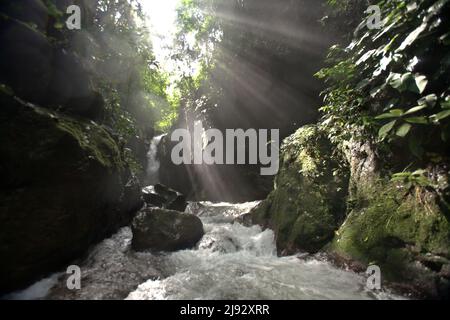 Lichtstrahlen fallen auf den Bach und die Umgebung unterhalb des Pendung Semurup Wasserfalls in der Nähe des Pendung Mudik Dorfes in Air Hangat, Kerinci, Jambi, Indonesien. Stockfoto