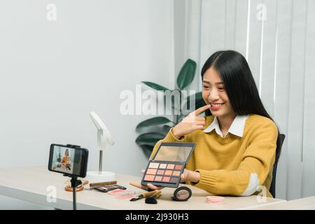 Junge asiatische Frau vloggt sich über Beauty-Produkte mit einem Video für ihren Blog über Kosmetik mit Handy-Kamera zu Hause machen. Stockfoto