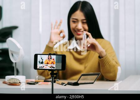 Junge asiatische Frau vloggt sich über Beauty-Produkte mit einem Video für ihren Blog über Kosmetik mit Handy-Kamera zu Hause machen. Stockfoto