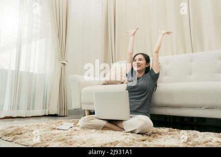 Asiatische Frau wirft Papiere im Wohnzimmer, als sie ihre Arbeit mit ihrem Laptop auf ihrem Schoß beendet. Stockfoto