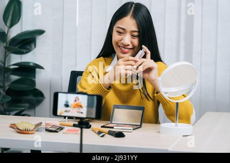 Junge asiatische Frau vloggt sich über Beauty-Produkte mit einem Video für ihren Blog über Kosmetik mit Handy-Kamera zu Hause machen. Stockfoto