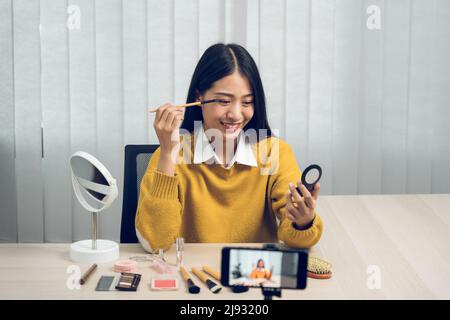 Junge asiatische Frau vloggt sich über Beauty-Produkte mit einem Video für ihren Blog über Kosmetik mit Handy-Kamera zu Hause machen. Stockfoto