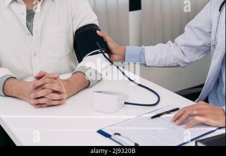 Der asiatische Arzt verwendet zum Zeitpunkt seiner jährlichen Untersuchung den Blutdruck-Monitor eines Patienten und erklärt seinen Blutdruck. Stockfoto