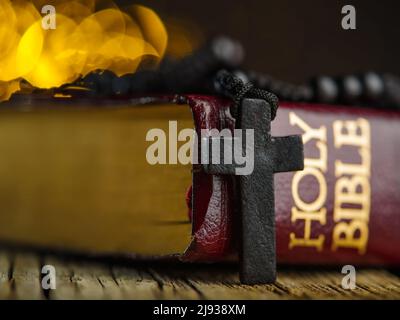 Makroaufnahme. Das Buch der Heiligen Bibel und das katholische Kreuz auf dem Rosenkranz auf einem Holztisch auf gelbem Hintergrund mit funkelnden Lichtern. Symbole von Stockfoto