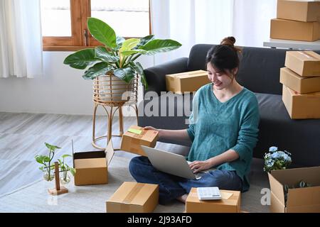 Lächelnde junge Frau, die ihren Laptop benutzt und Paketboxen für die Lieferung an die Kunden vorbereitet. E-Commerce, Online-Verkaufskonzept Stockfoto